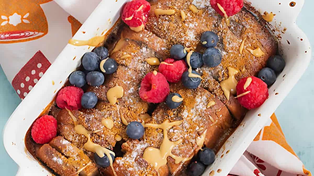 A baked dish with golden-brown French toast topped with fresh raspberries, blueberries, powdered sugar, and a drizzle of peanut butter. It sits on a colorful cloth with orange and red patterns.