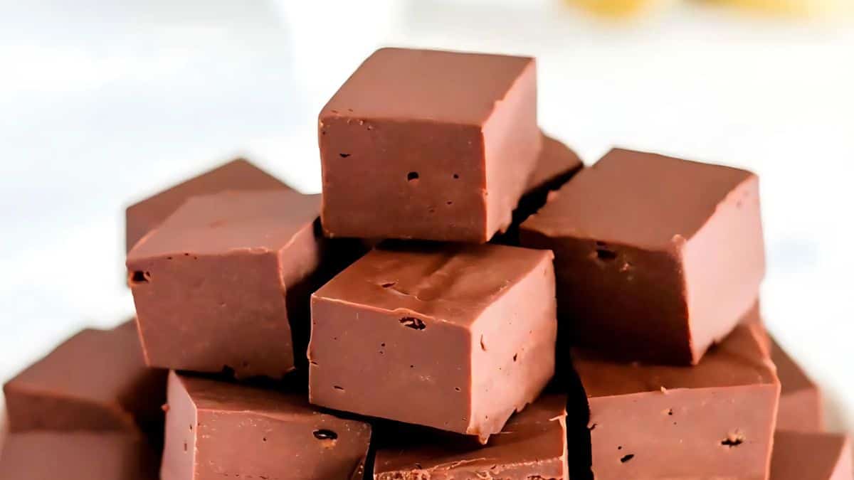A stack of smooth chocolate fudge cubes, each uniformly shaped, on a light background. The rich, glossy texture and dark brown color of the fudge are prominent, hinting at its creamy and indulgent nature.