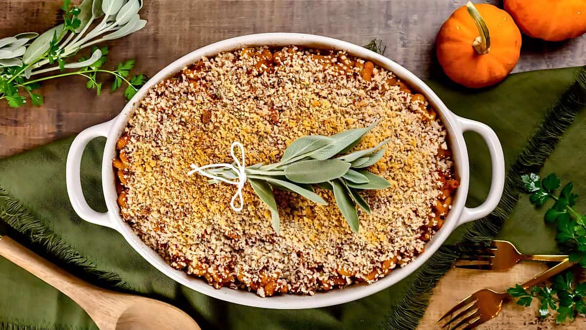 A casserole dish filled with a breadcrumb-topped baked dish garnished with fresh sage. It’s placed on a green cloth, surrounded by small pumpkins, a wooden spoon, and gold forks on a wooden table.