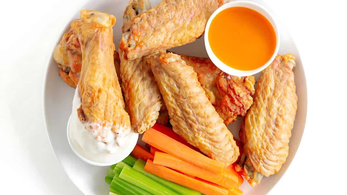 A plate of crispy chicken wings with two dipping sauces, one white and one orange. The plate also includes fresh celery and carrot sticks, all set against a white background.
