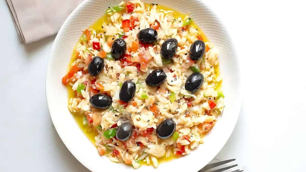 A colorful dish with rice, chopped red and green bell peppers, topped with shiny black olives, served on a white plate. Silverware and a napkin are visible beside the plate on a light surface.