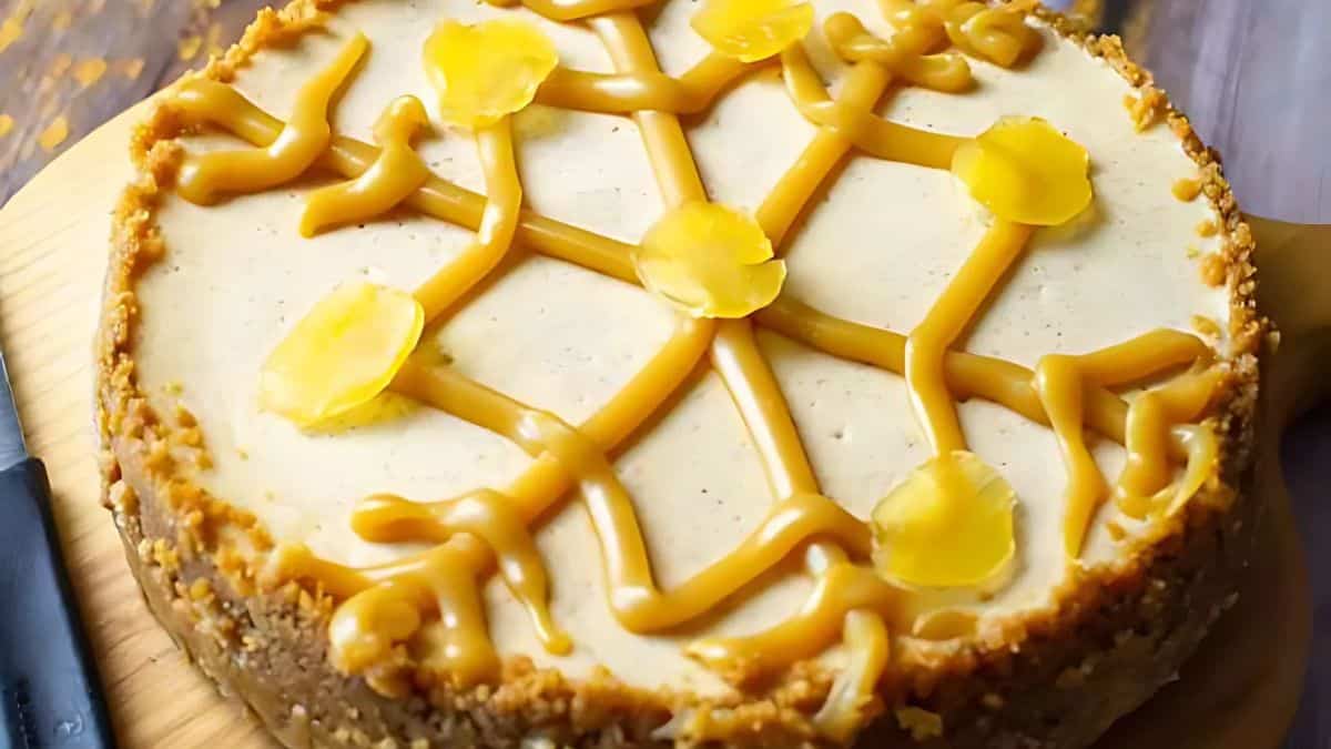A round cheesecake with a nutty crust, topped with caramel drizzle and yellow flower-like decorations, on a wooden board.