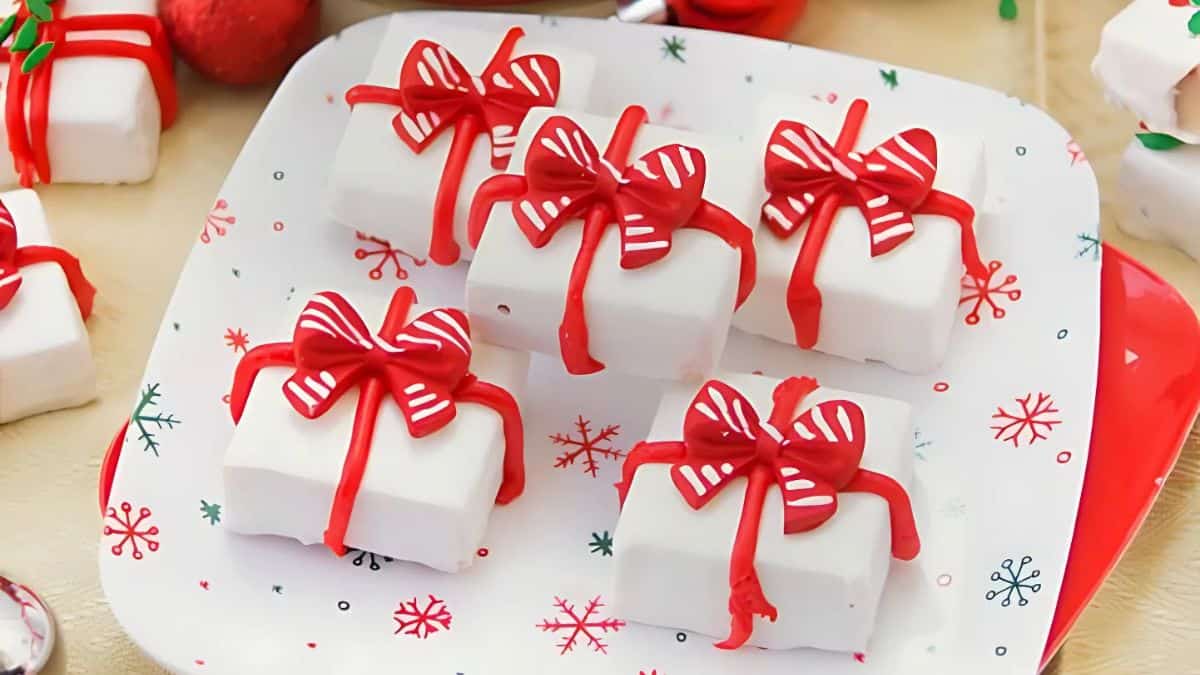 A plate with four white square cakes, each topped with a red ribbon designed like a gift. The plate is decorated with red and green snowflakes, capturing a festive holiday theme.