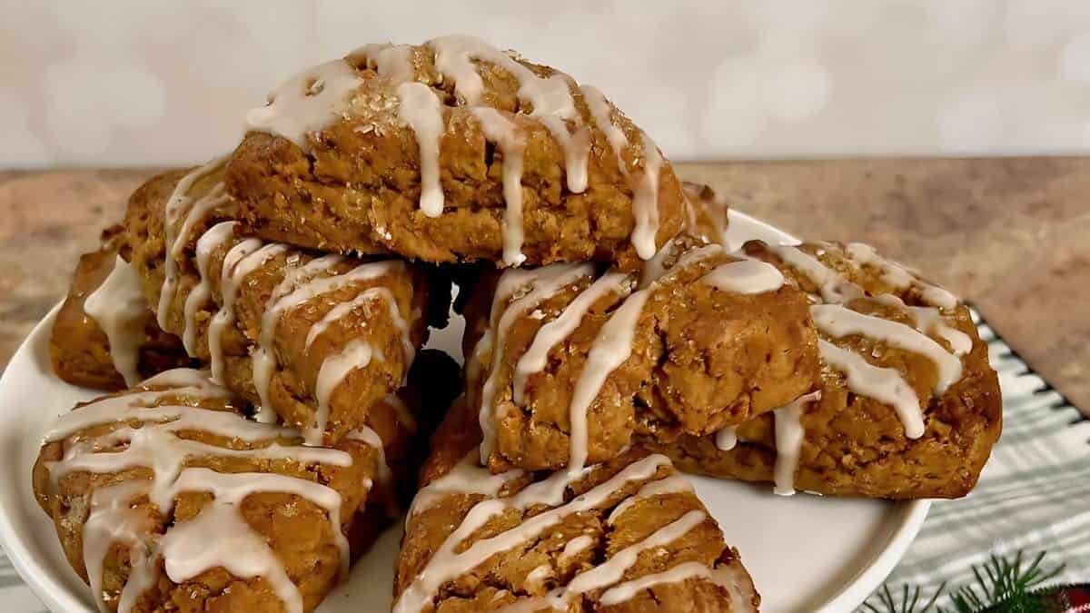 A stack of pumpkin scones, drizzled with icing, is presented on a white plate. The scones are golden brown with a glossy glaze, set against a soft-focus kitchen background.