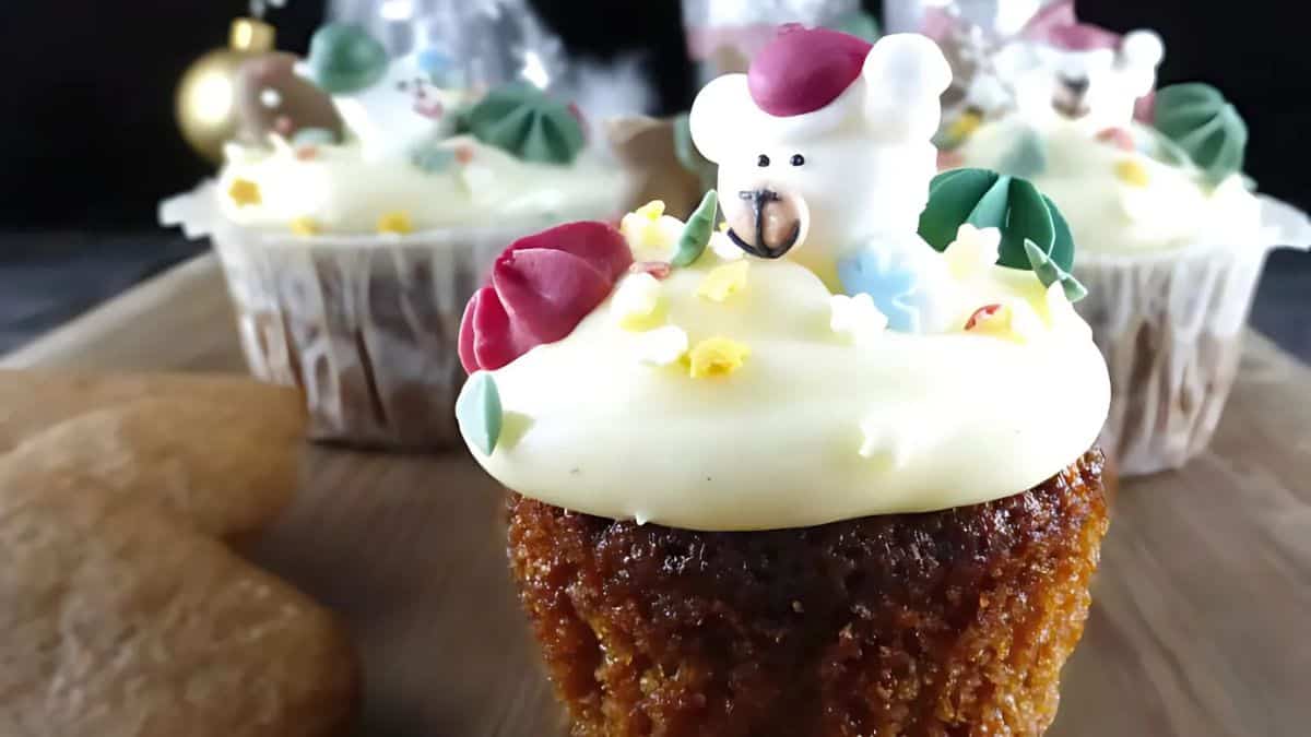 A close-up of a festive cupcake topped with white frosting and a small bear-shaped decoration. The cupcake is surrounded by more cupcakes with similar decorations and two heart-shaped cookies on a wooden board.