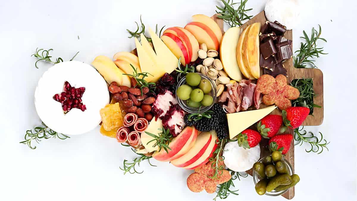 A wooden board artfully arranged with assorted cheeses, fruits, nuts, chocolates, olives, and cured meats. Garnished with rosemary sprigs, the board features strawberries, apple slices, pistachios, and decorative crackers.