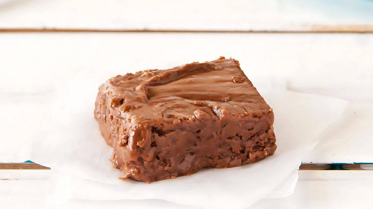 A piece of chocolate fudge sits on a white piece of parchment paper, showcasing its rich, smooth texture and glossy finish. The fudge is square-shaped and placed on a light-colored wooden surface.