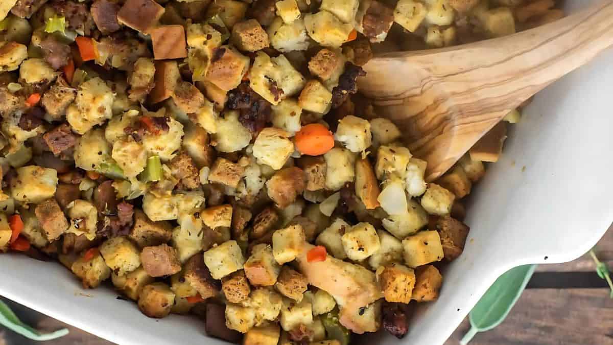 A wooden spoon rests in a baking dish filled with golden-brown stuffing. The stuffing includes toasted bread cubes, chopped carrots, celery, and herbs. Steam rises from the dish, indicating it's freshly baked and hot.