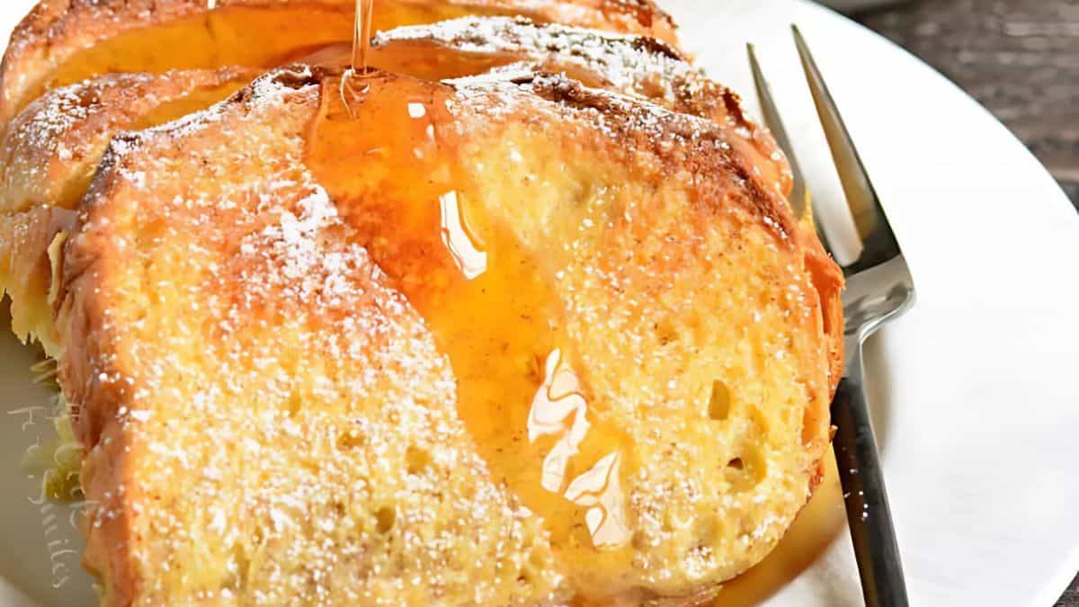 Close-up of French toast on a white plate, drizzled with syrup and dusted with powdered sugar. A fork rests beside the toast, and the background features a rustic wooden table.