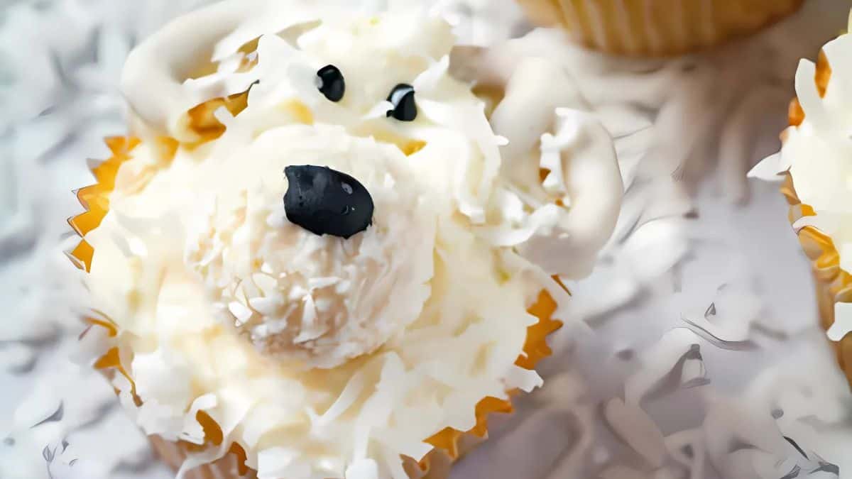 A cupcake decorated to resemble a polar bear, with white frosting, coconut flakes for fur, a candy nose, and small black eyes. The background is scattered with more coconut flakes.