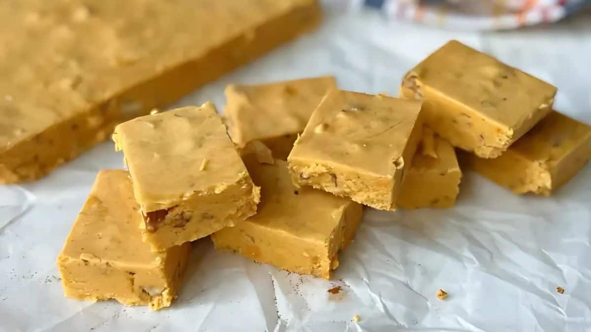 Squares of creamy fudge are stacked on a crumpled white parchment paper. The fudge has a smooth, tan appearance with small bits visible inside, likely indicating nuts or caramel. A larger uncut slab of fudge is in the background.