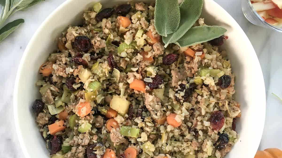 A bowl filled with a savory dish consisting of diced vegetables, rice, cranberries, and ground meat, garnished with fresh sage leaves. The ingredients are mixed together and attractively presented on a white marble surface.