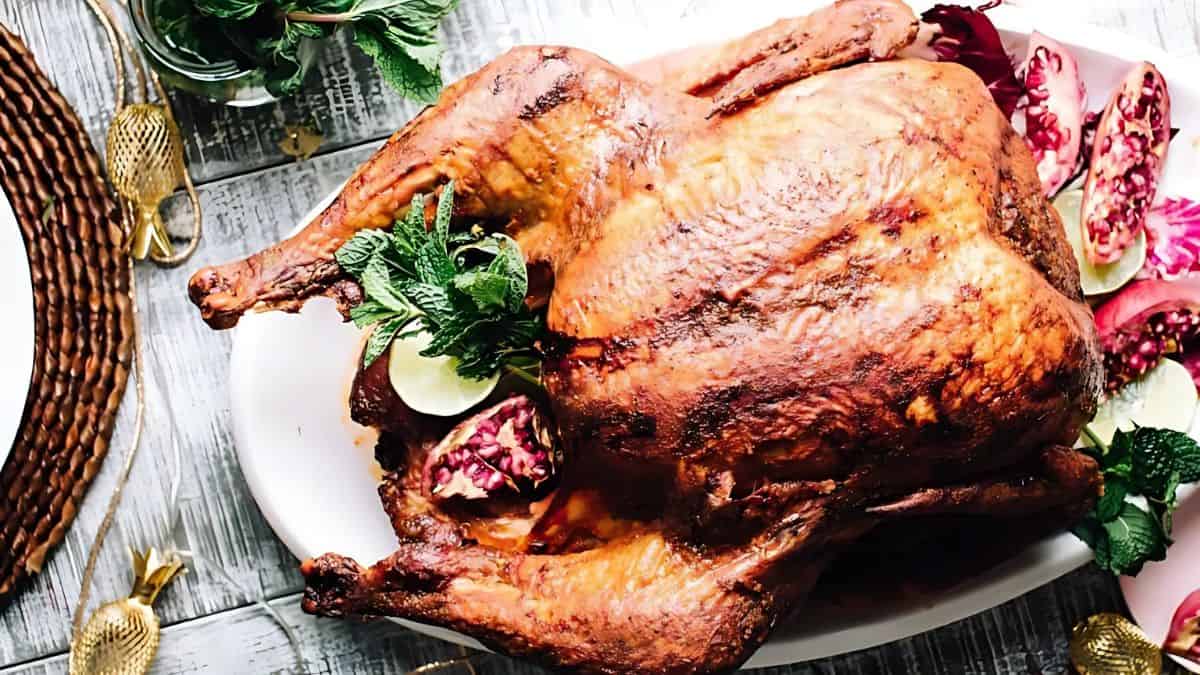 A roasted turkey on a white platter, garnished with lime slices, pomegranate seeds, and fresh mint. The table is set with a brown woven placemat and gold-colored utensils.