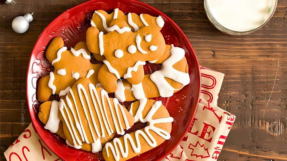 A red plate filled with frosted gingerbread cookies shaped like people and trees is displayed on a wooden table. Next to the plate is a glass of milk. Holiday-themed napkins are partially visible underneath.