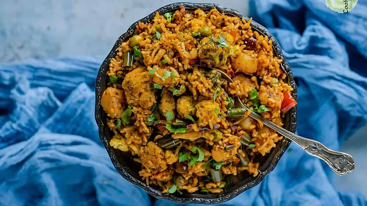 A black bowl filled with spicy mixed vegetable biryani, garnished with fresh herbs. The dish features rice combined with assorted vegetables and spices. A silver fork rests on the side. A blue fabric is draped underneath the bowl.