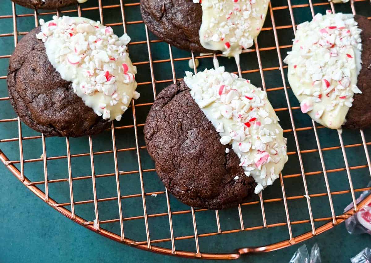 Chocolate cookies partially dipped in white chocolate, topped with crushed peppermint pieces, resting on a copper cooling rack.