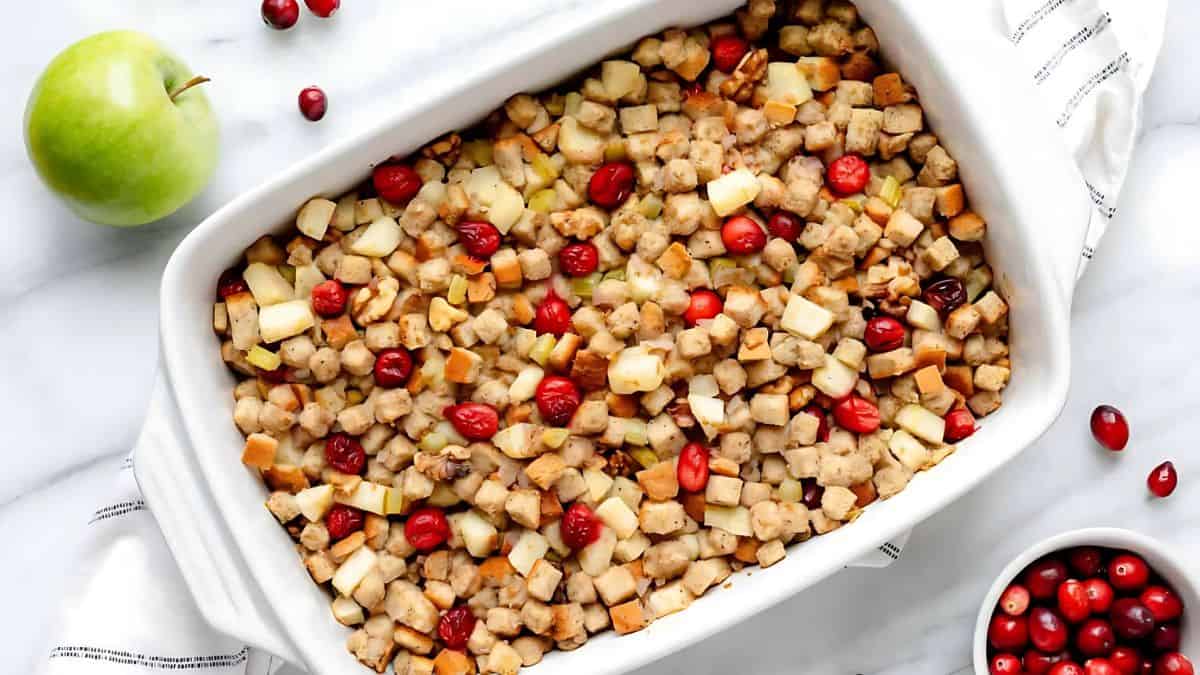 A white casserole dish filled with bread cubes, sausage pieces, cranberries, and apple chunks on a marble surface. A green apple and scattered cranberries are beside the dish, along with a small bowl of cranberries.