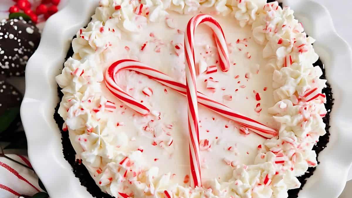 A festive dessert featuring a creamy white filling topped with crushed peppermint candy and two whole candy canes arranged in an X shape, set in a chocolate crust with a scalloped edge, surrounded by dollops of whipped cream.