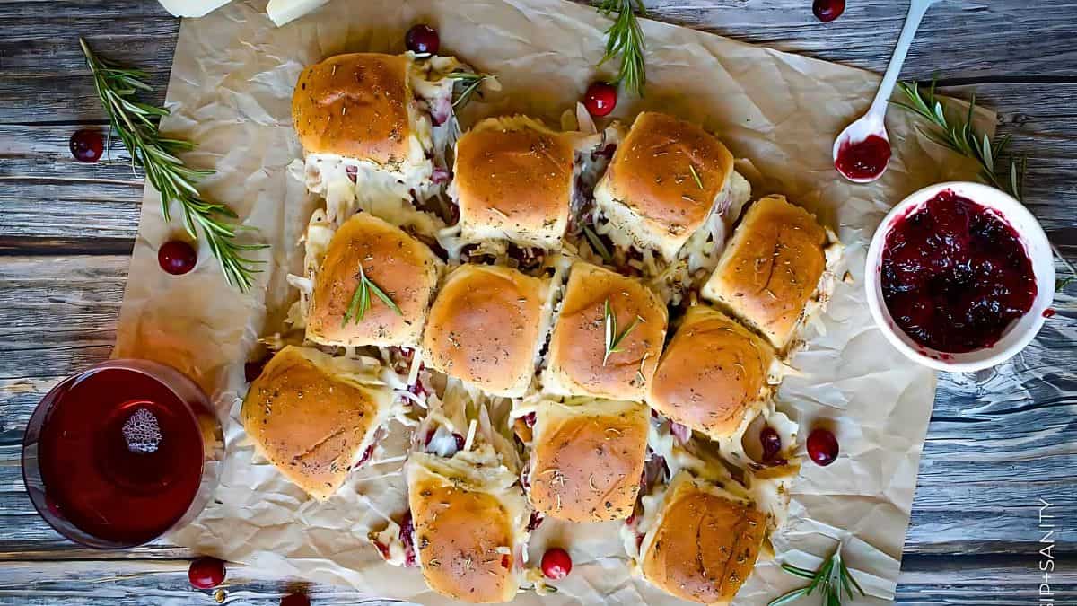 A batch of slider sandwiches on parchment paper, topped with melted cheese and herbs. Surrounded by sprigs of rosemary, fresh cranberries, a bowl of cranberry sauce, and a glass of red drink on a wooden surface.