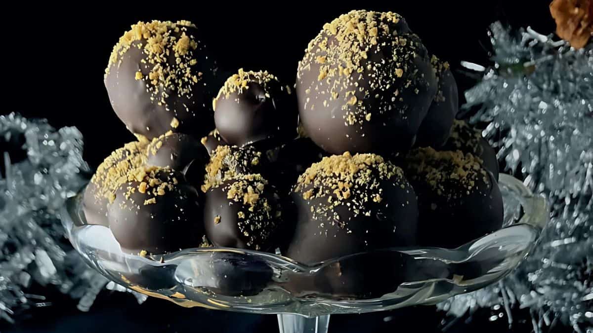 A glass plate holds a stack of chocolate truffles, each coated with a layer of dark chocolate and sprinkled with crushed nuts. Silver tinsel is blurred in the background, giving a festive touch to the dark, elegant setting.