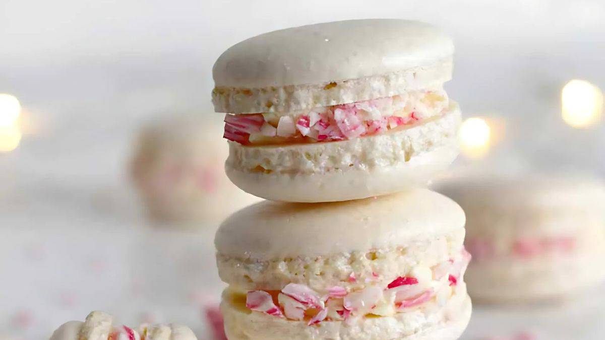 Two white macarons stacked on top of each other, filled with cream and crushed peppermint candy, with a soft-focus background featuring more macarons and subtle twinkle lights.