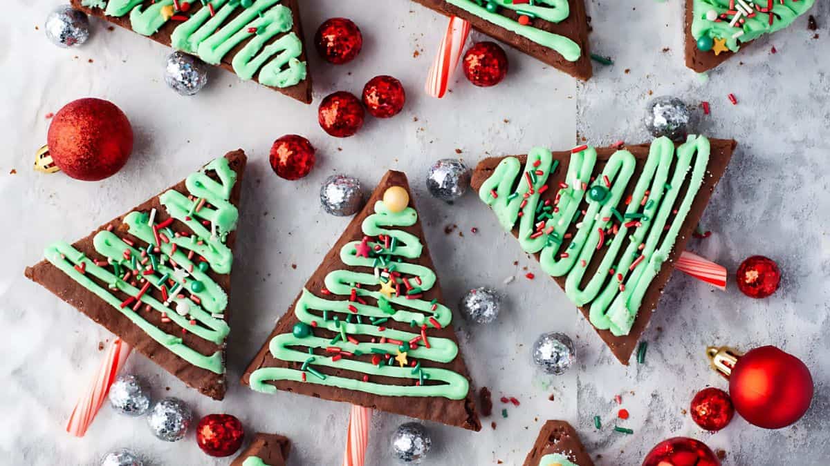 Triangular chocolate cookies decorated as Christmas trees with green icing and sprinkles, each on peppermint stick trunks. Surrounded by red and silver ornaments on a white surface.