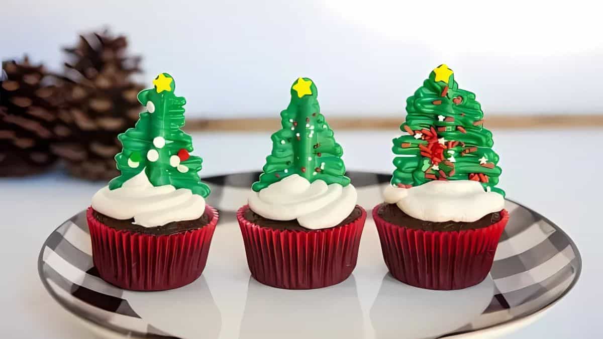 Three festive cupcakes with red liners topped with white frosting and decorated with green Christmas tree toppers. Each tree features a yellow star and colorful sprinkles. Background includes a blurred pine cone.