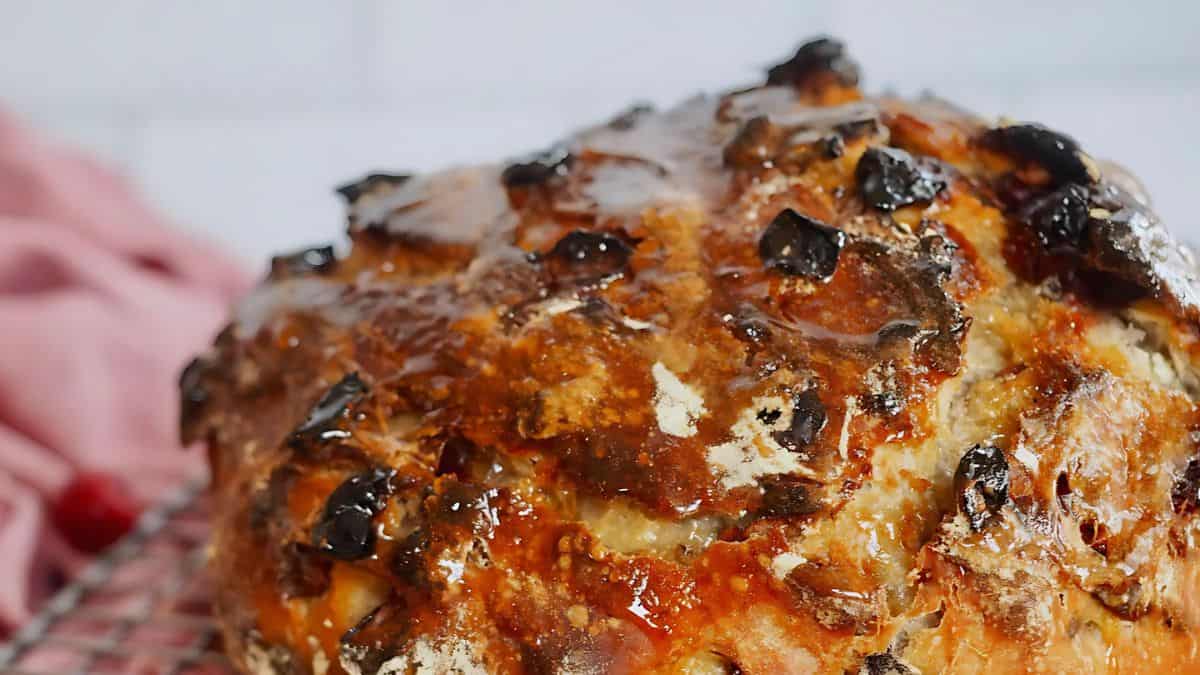 A close-up of a rustic, freshly baked loaf of bread with a golden crust, speckled with dark, baked olives. The bread sits on a cooling rack, with a soft pink cloth blurred in the background.