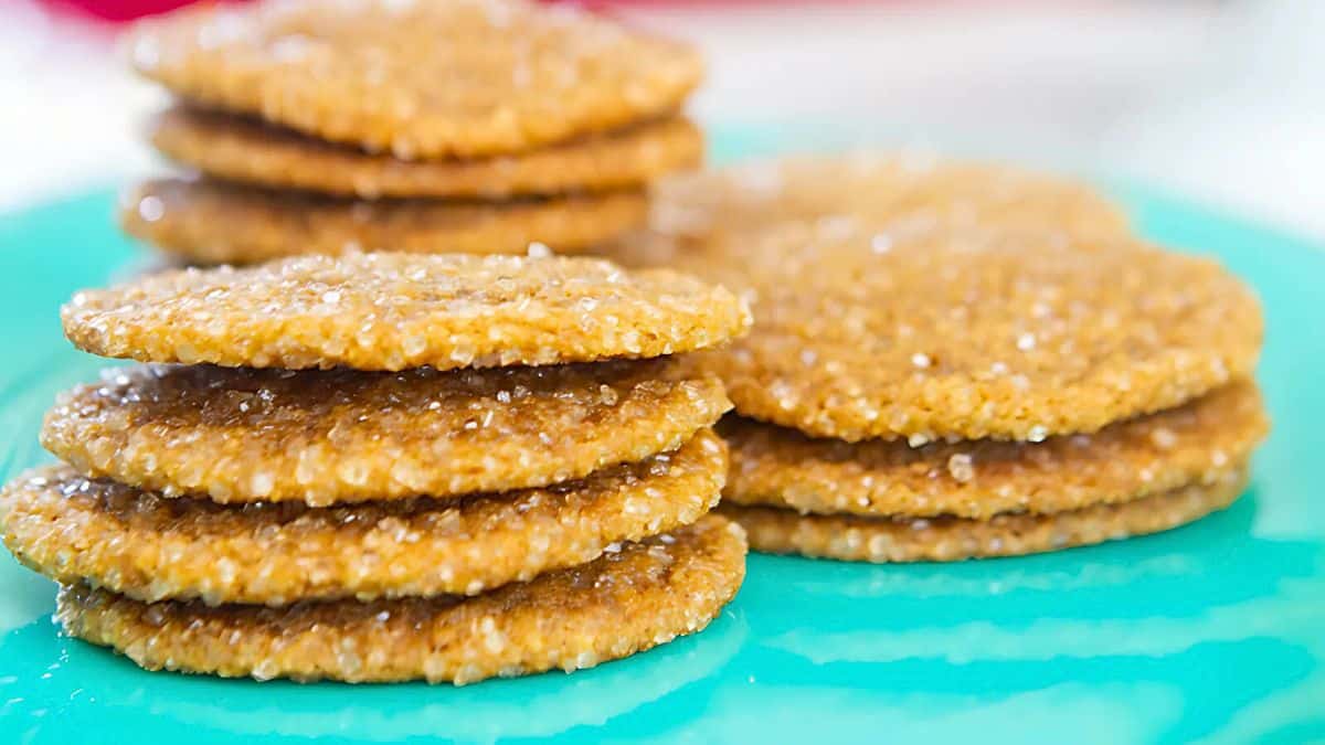A stack of thin, golden-brown cookies with sugar crystals on top. They are arranged on a bright turquoise plate. The background is slightly blurred, highlighting the texture and shimmer of the cookies.