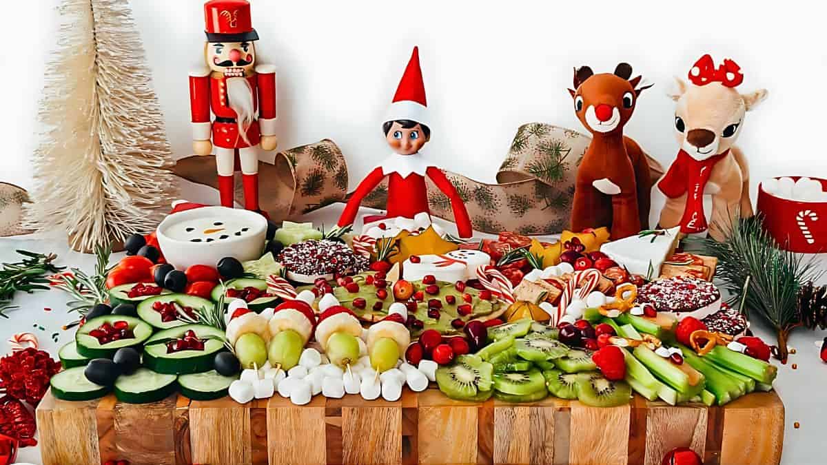 A festive charcuterie board features a colorful assortment of fruits, vegetables, and sweets. It is decorated with holiday-themed toys, including a nutcracker, an elf, and two reindeer figurines, set against a wintery backdrop with a pine tree.