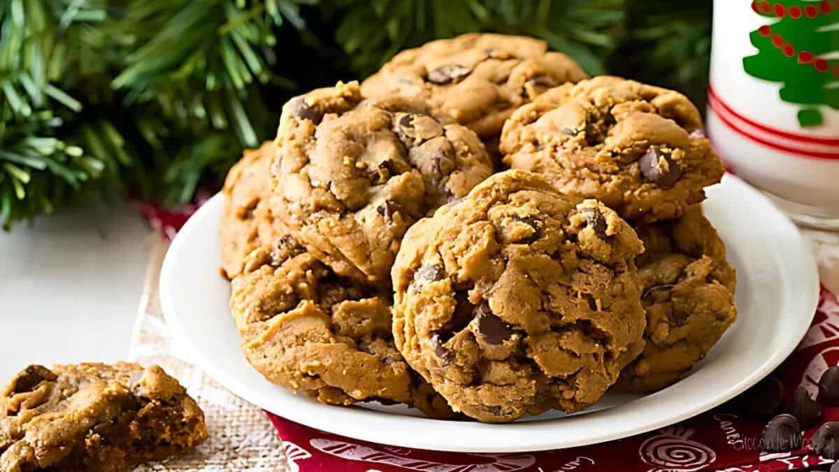 A plate of chocolate chip cookies is set on a table next to a glass of milk. The backdrop includes festive holiday decorations and a green pine tree branch, creating a cozy, holiday atmosphere.
