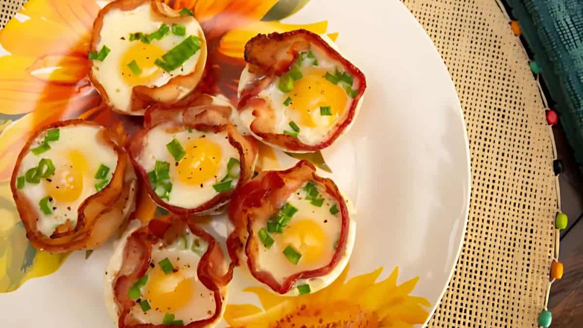 A plate with six bacon-wrapped egg cups garnished with chopped green onions, resting on a floral-patterned plate atop a woven placemat.