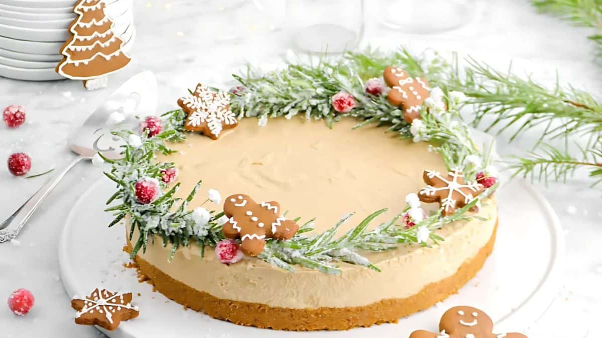 A festive cheesecake topped with a wreath of evergreen sprigs, sugared cranberries, and gingerbread cookies, including snowflakes and gingerbread people. The scene is set on a snowy white background with additional gingerbread cookies nearby.