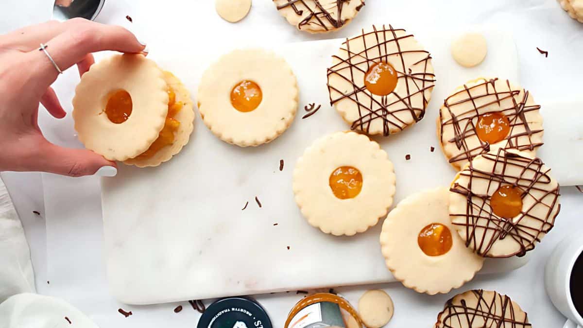 A hand reaches for a stack of round sandwich cookies filled with orange jam on a white marble surface. Some cookies are drizzled with chocolate, and small containers of jam are visible nearby.