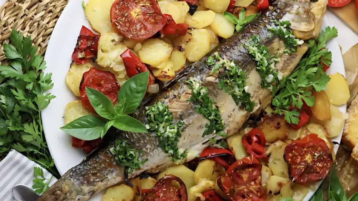 Grilled fish garnished with herbs, surrounded by sliced potatoes, tomatoes, and red peppers on a white plate. Fresh basil and parsley add a touch of green. The dish is set on a wicker surface.