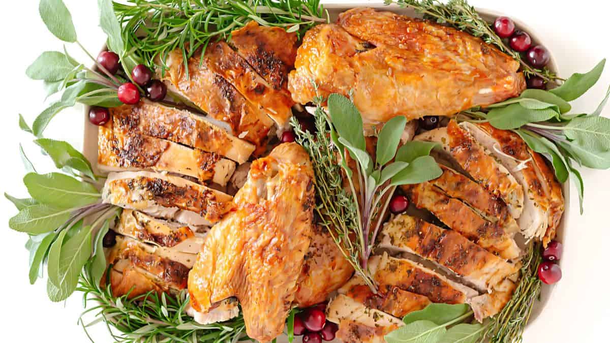 Sliced roasted turkey breast garnished with fresh rosemary, sage, thyme, and cranberries arranged on a white platter.