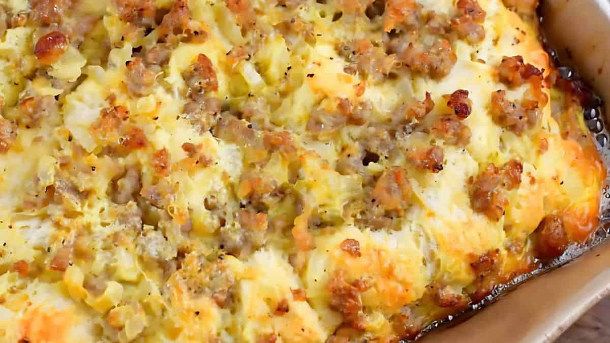 A close-up of a savory casserole topped with golden-brown melted cheese and browned sausage. The dish appears hot and freshly baked, with visible bits of sausage and cheese on the surface.