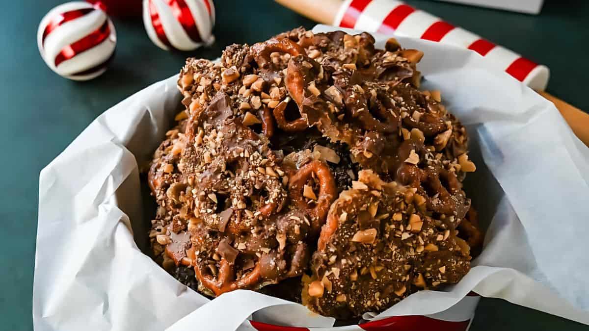 A bowl of pretzel toffee clusters is set on a dark surface. The clusters are topped with crushed nuts and chocolate. In the background, there are candy cane ornaments and a red and white striped object.
