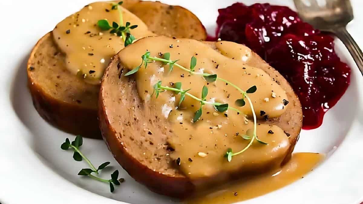 Two slices of vegan turkey roast topped with gravy and garnished with fresh herbs, served on a white plate. Cranberry sauce accompanies the dish on the side.