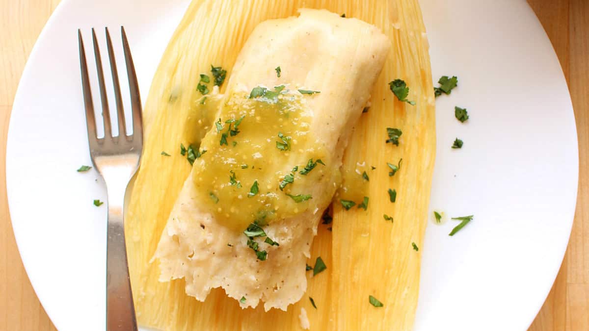 A Vegetable tamale topped with green salsa and garnished with fresh cilantro sits on a corn husk, placed on a white plate beside a fork.