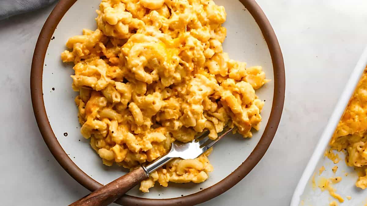 A plate of creamy macaroni and cheese sits on a table. The mac and cheese is golden with a slightly browned top, served on a round white plate with a brown rim. A fork rests on the plate, ready to dig in.