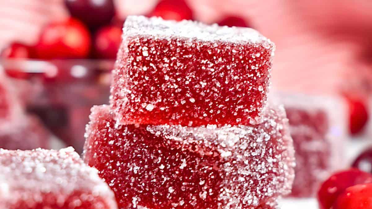 Close-up of sugar-coated red jelly cubes stacked in a pile. The background features blurred red and white elements, including glimpses of cherries or similar fruit.