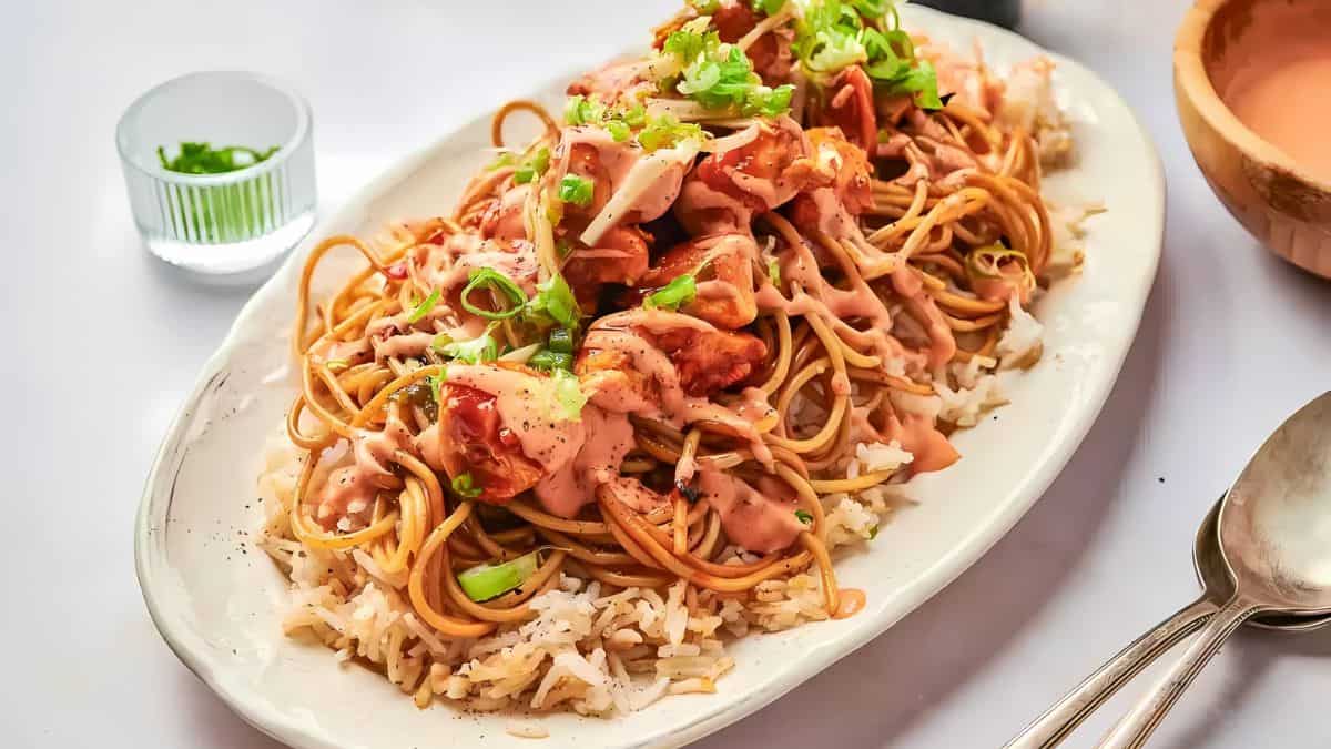 A plate of spaghetti served over rice, topped with pieces of chicken, drizzled with a creamy sauce, and garnished with chopped green onions. A small glass bowl with more green onions is in the background.