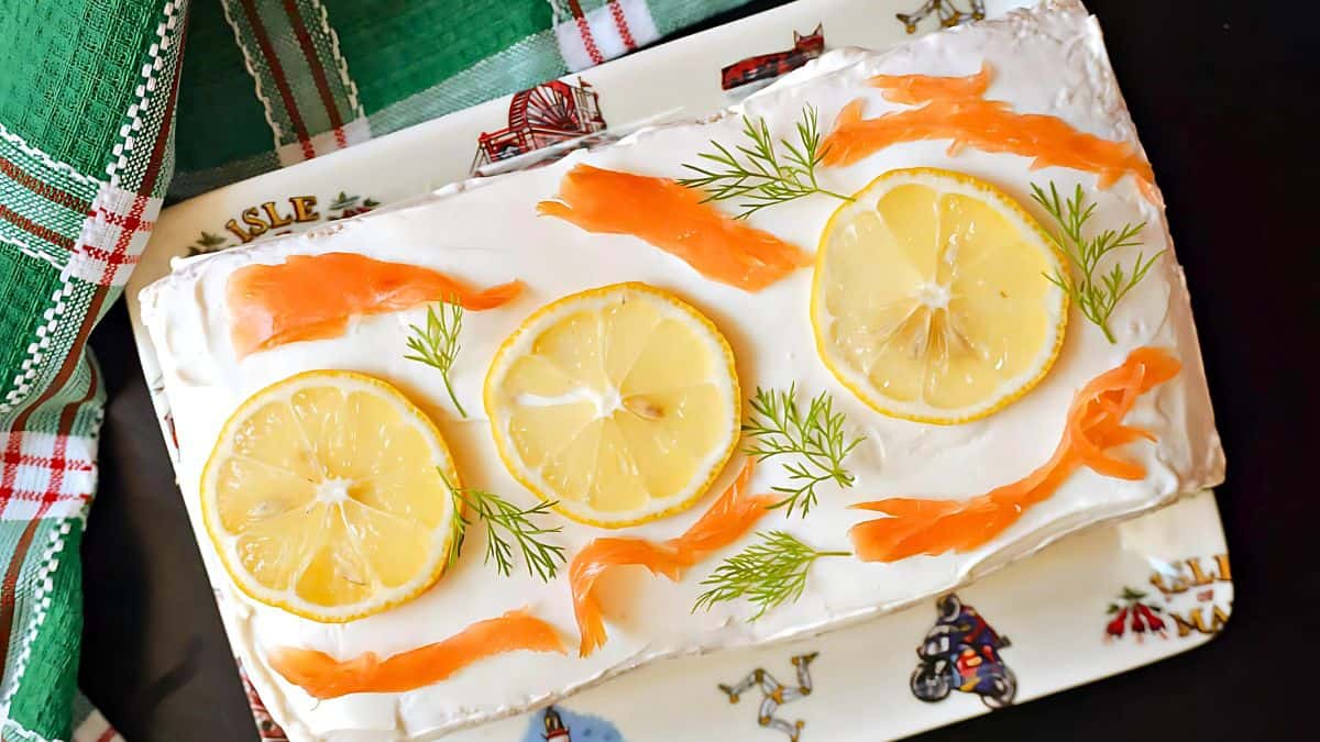 A rectangular cake topped with three slices of lemon, thin strips of smoked salmon, and sprigs of dill. It's served on a decorative plate with a green and red checkered cloth beside it.