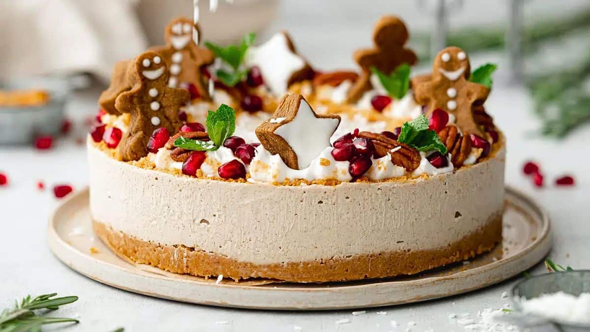 A festive cake topped with gingerbread cookies, white star-shaped decorations, pomegranate seeds, pecans, and mint leaves. The cake is set on a light-colored round platter, creating a cheerful holiday presentation.