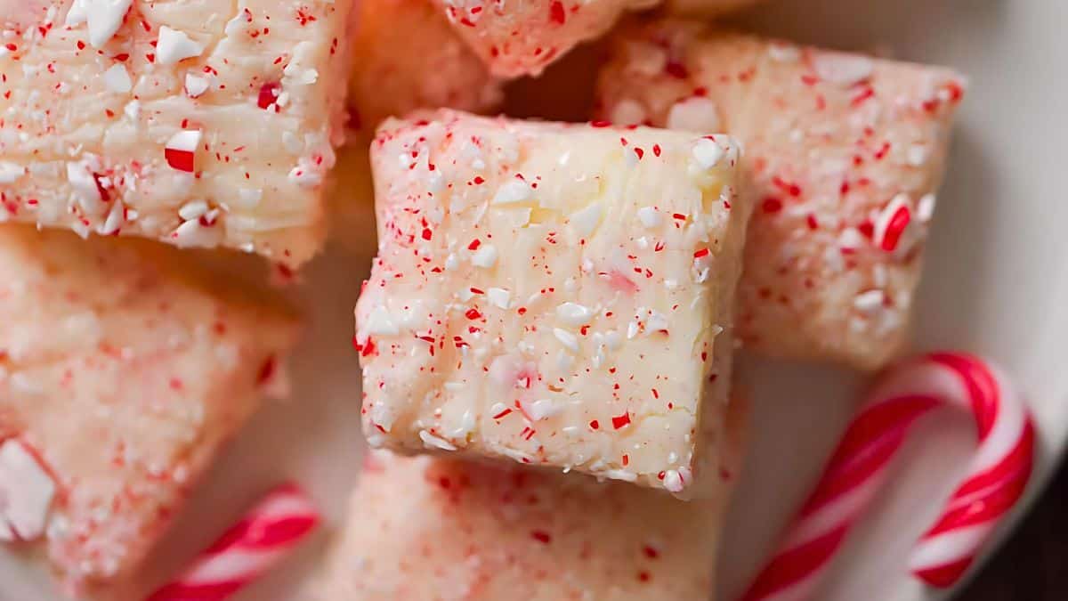 Close-up of peppermint bark fudge squares sprinkled with crushed candy canes, arranged on a plate. Candy canes are visible in the background, adding a festive and sweet holiday touch.