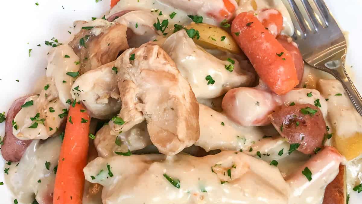 A plate of creamy chicken served with baby carrots and roasted potatoes. The dish is garnished with chopped herbs, and a silver fork is placed on the side.