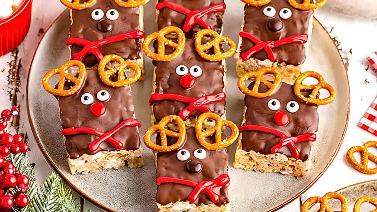 Festive reindeer-themed treats with chocolate-coated faces, pretzel antlers, candy eyes, and red noses, arranged on a round plate. Red frosting decorates each treat as a scarf. Holiday decorations surround the plate.