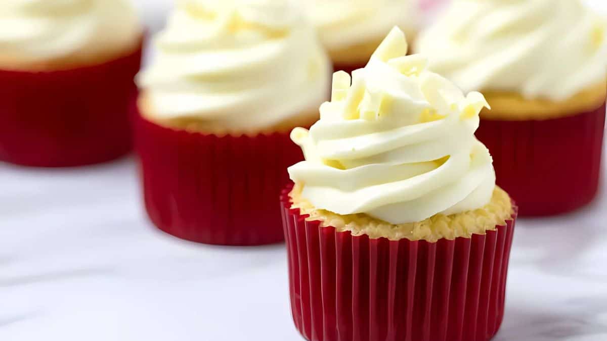Close-up of vanilla cupcakes topped with creamy white frosting, each in a red cupcake liner. They are arranged on a light surface, with some partially visible in the background.