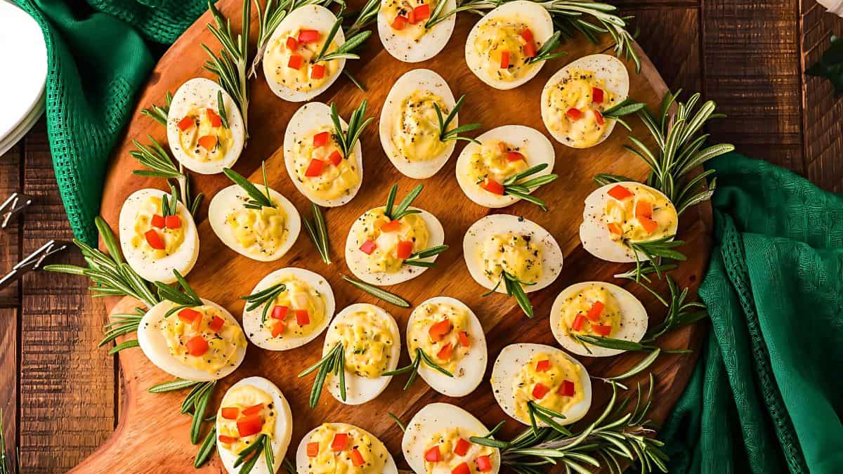 A platter of deviled eggs garnished with diced red peppers and rosemary sprigs, arranged in a circular pattern on a wooden board. A green cloth napkin is partially visible on the side.